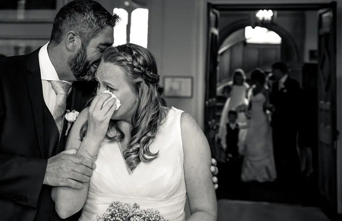 Black and white of the groom resting his head on the tearful bride as she dabs her eyes with a tissue.