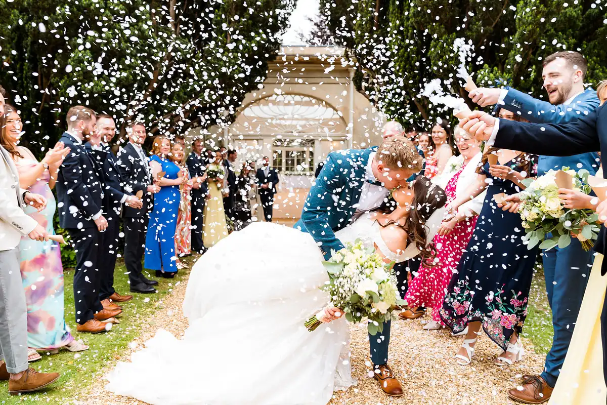 Groom dips the bride for a kiss as guests lining the path throw white confetti over them.