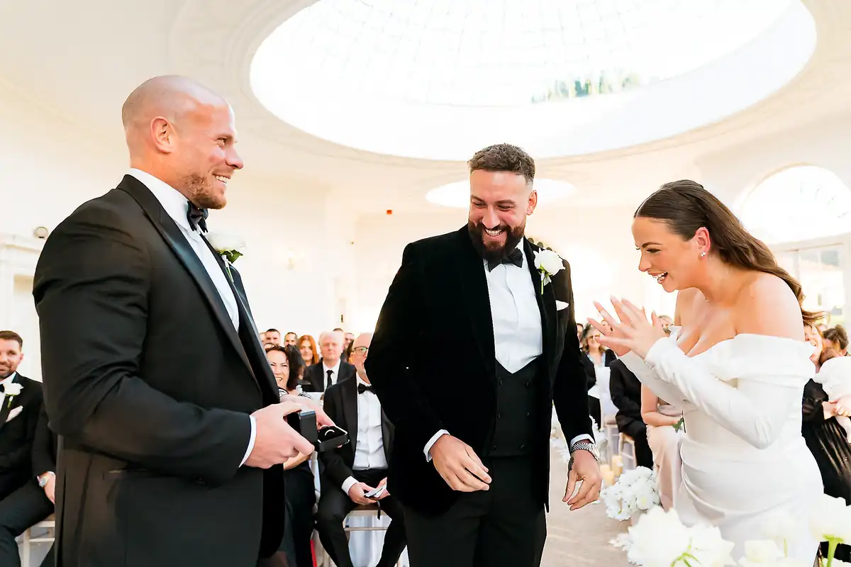 Groom laughing as the bride claps at the top of the aisle, best man grinning beside them under a round skylight.
