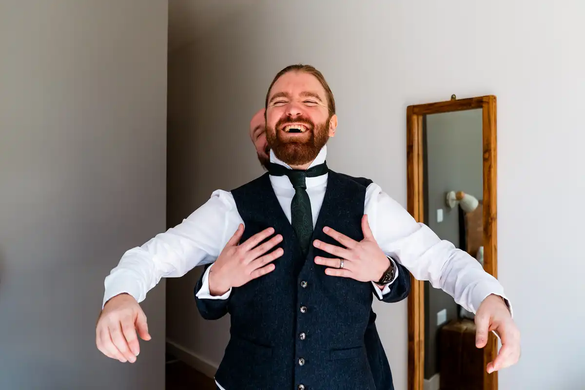 Groom laughing with his eyes shut as a friend behind him slips his arms through and pats his chest.