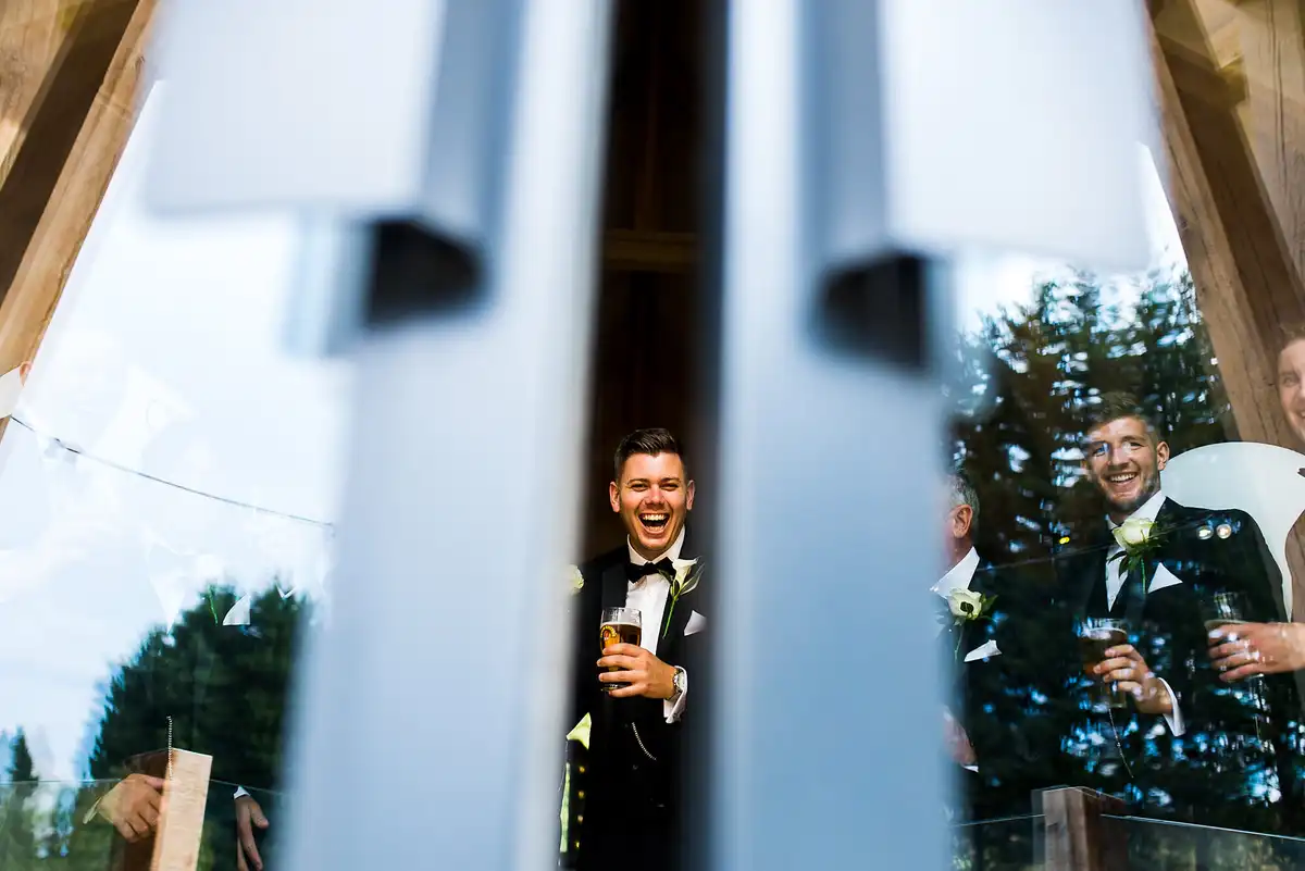 Groom in a bow tie laughing with a pint, seen through a gap in tall glass doors, a groomsman grinning on the right.