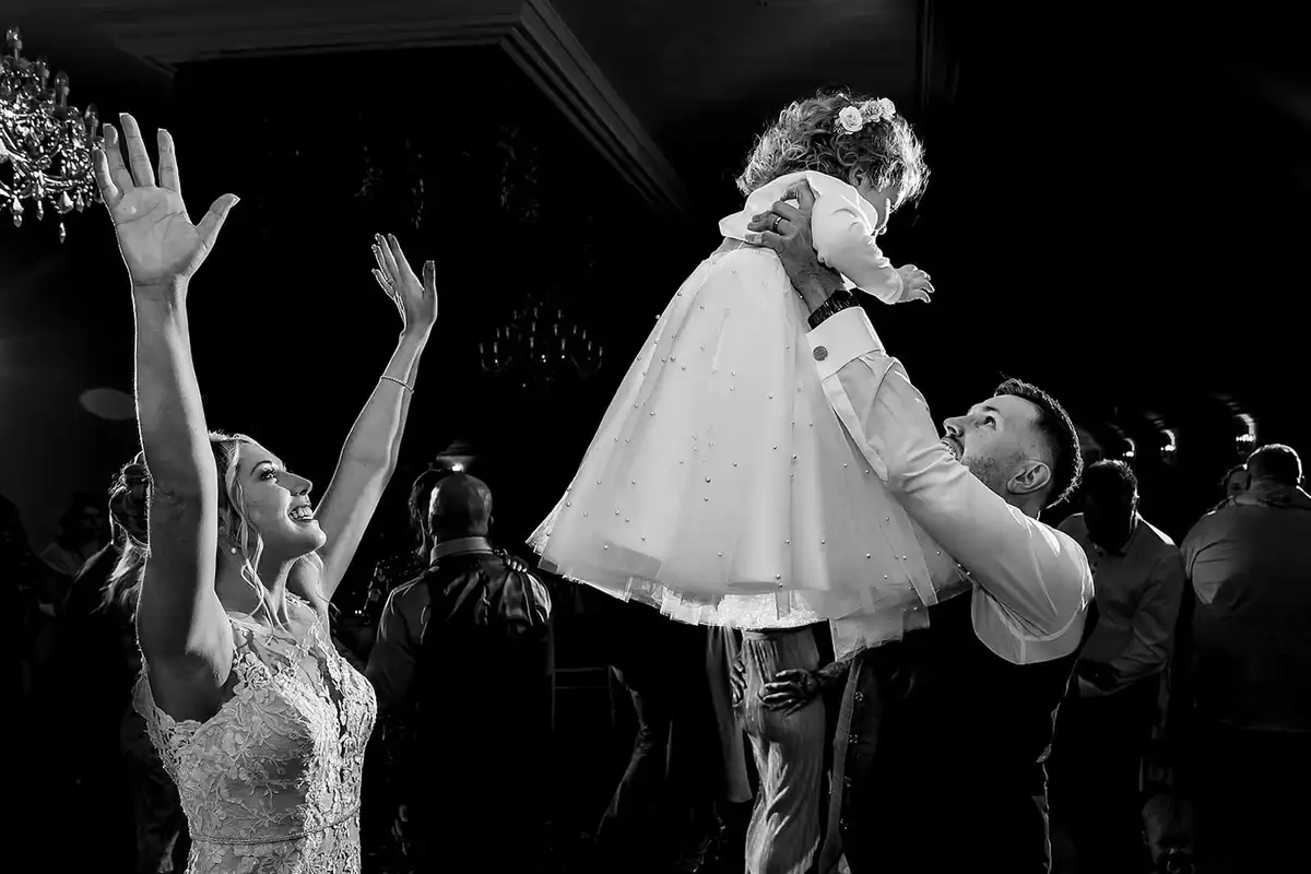 Black and white of a groom lifting a toddler high on the dance floor while the bride cheers, arms up.