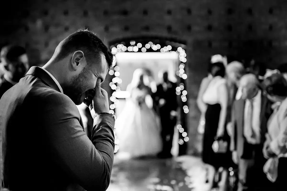 Black and white of the groom with his hand over his eyes as the bride appears in a doorway framed with fairy lights.