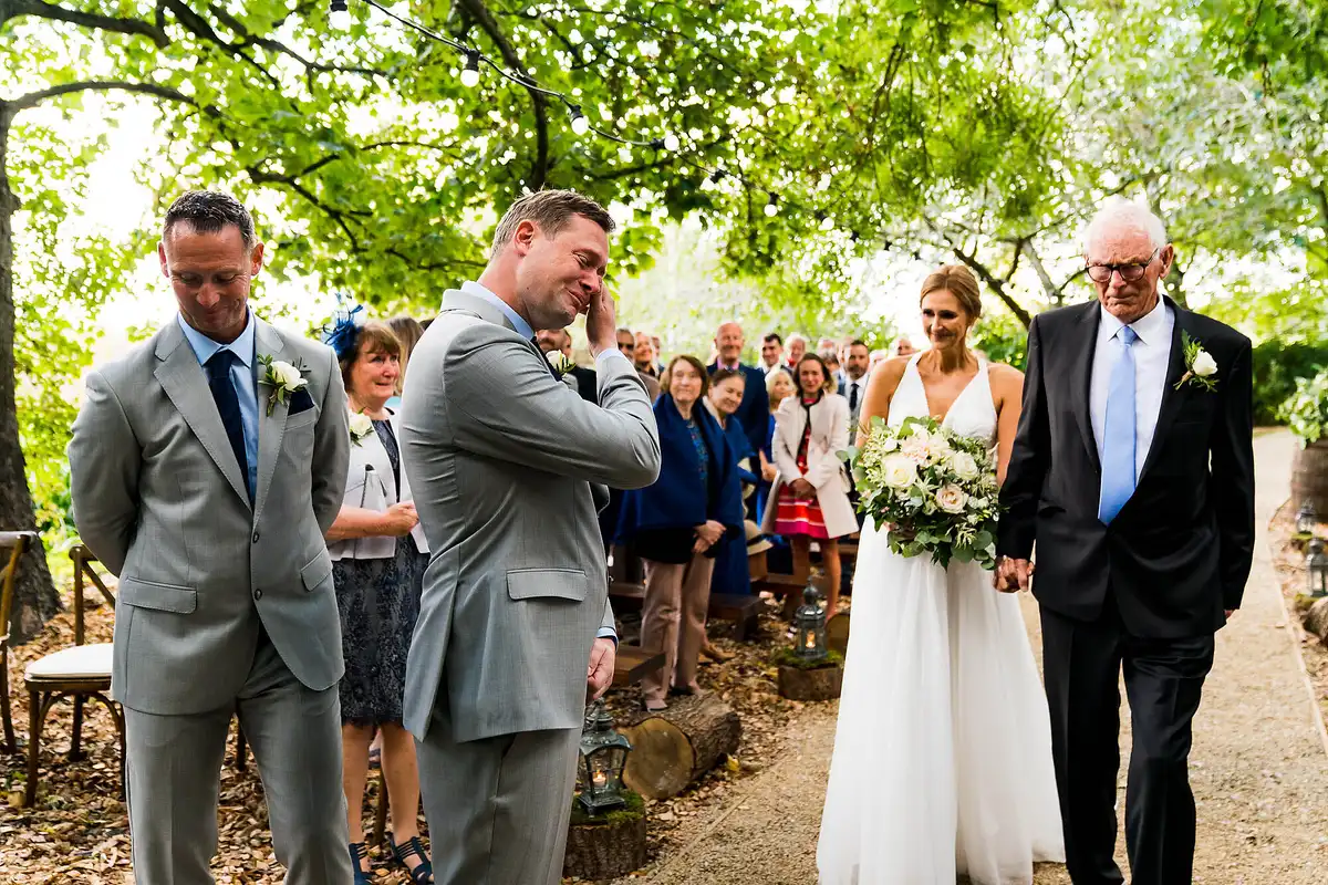 Groom wipes his eyes as the bride walks up a woodland aisle with an older man, guests standing under the trees.
