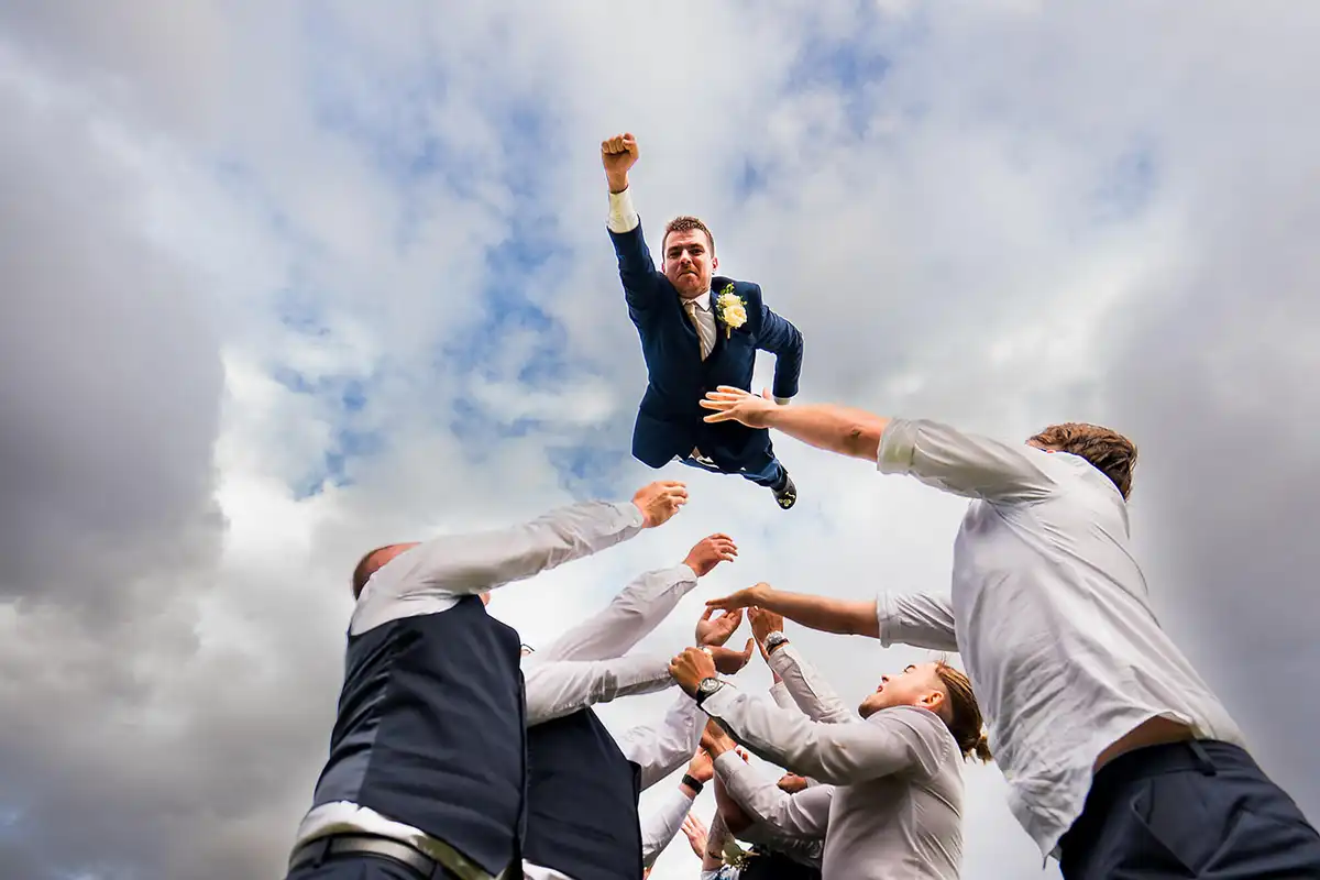 Groom thrown high into a cloudy sky by his groomsmen, one fist punched upwards like a superhero.