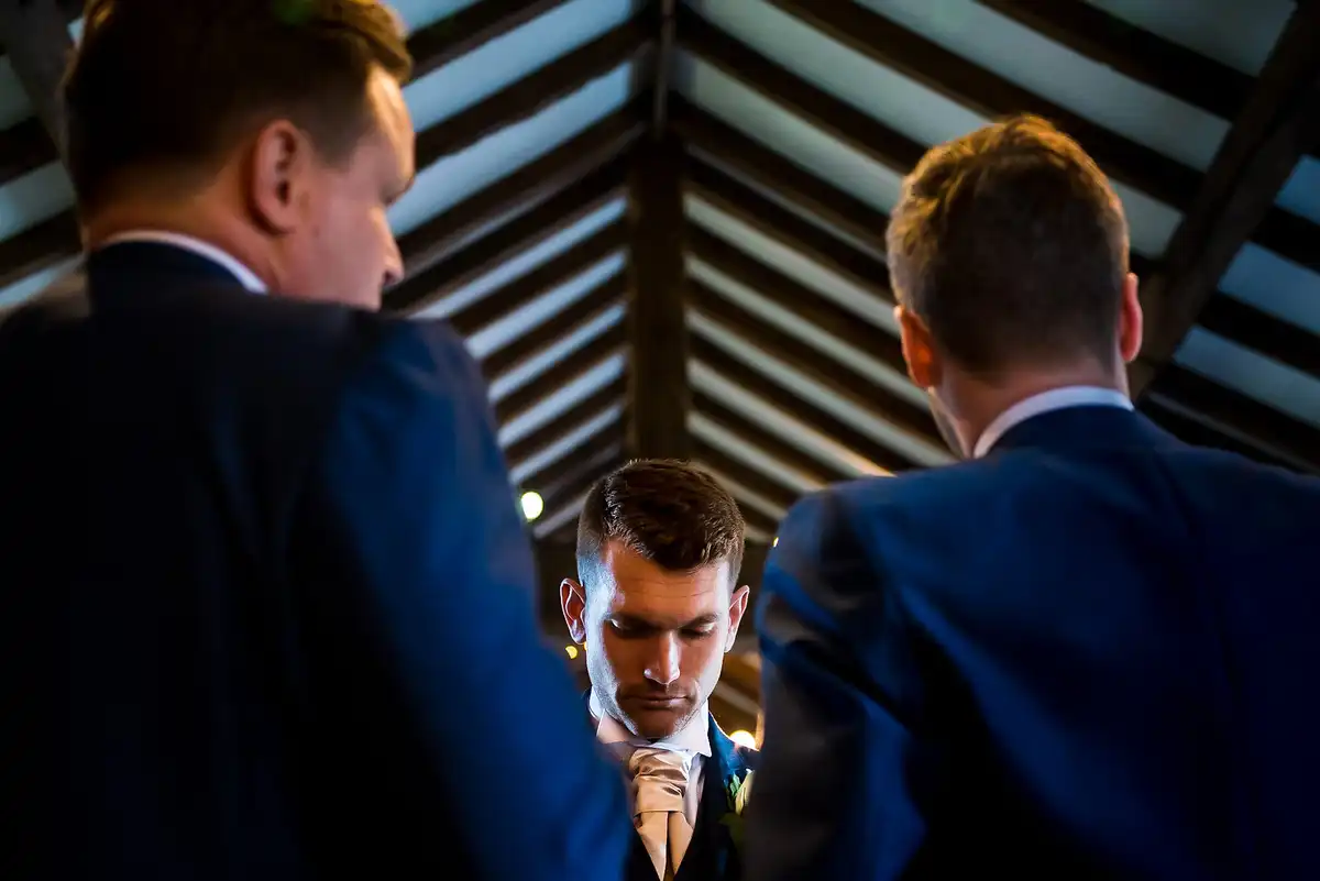 Groom looks down as two groomsmen lean in to sort his tie, seen from below under a timber roof.