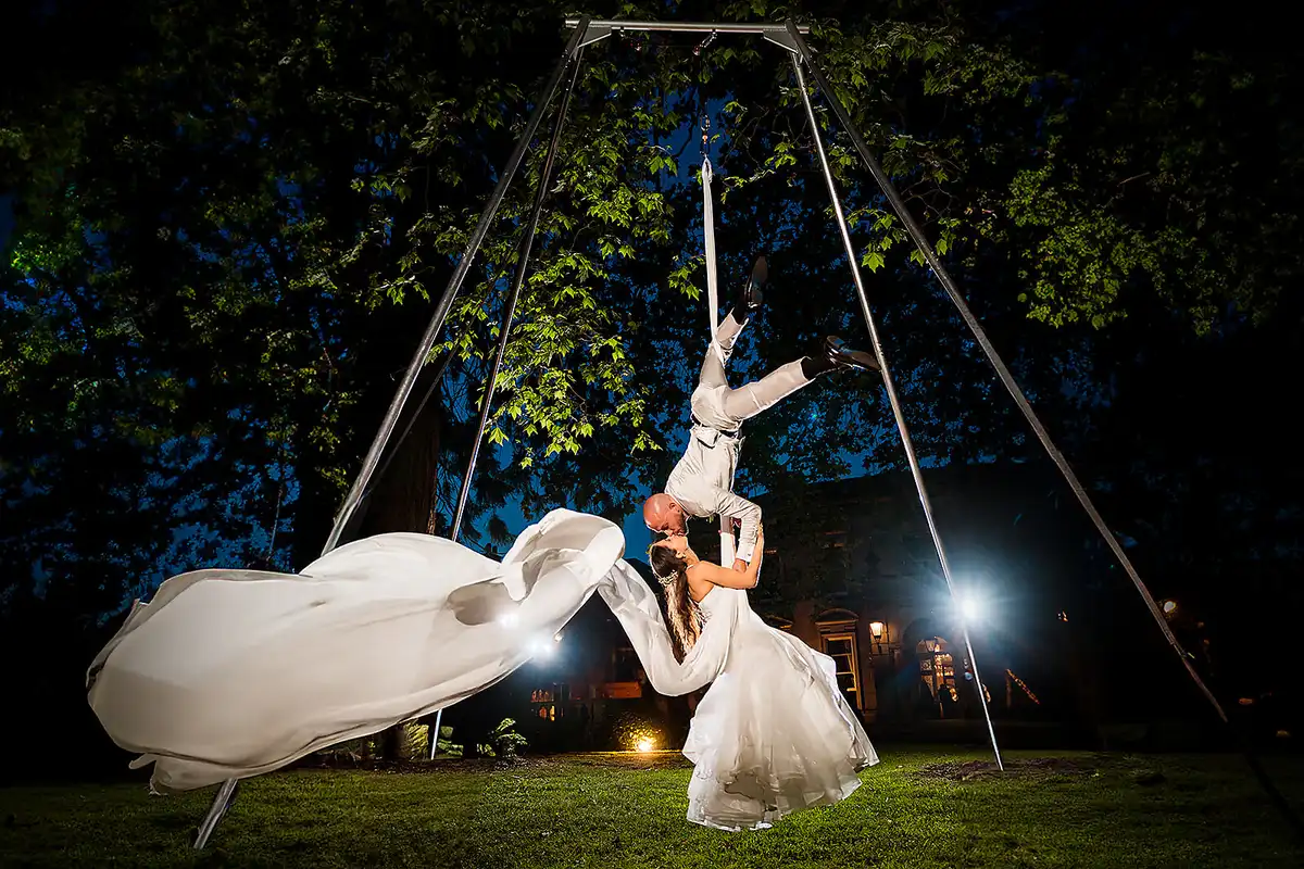 Groom hangs upside down from an aerial rig to kiss the bride at night, her long sheer cape flying out.