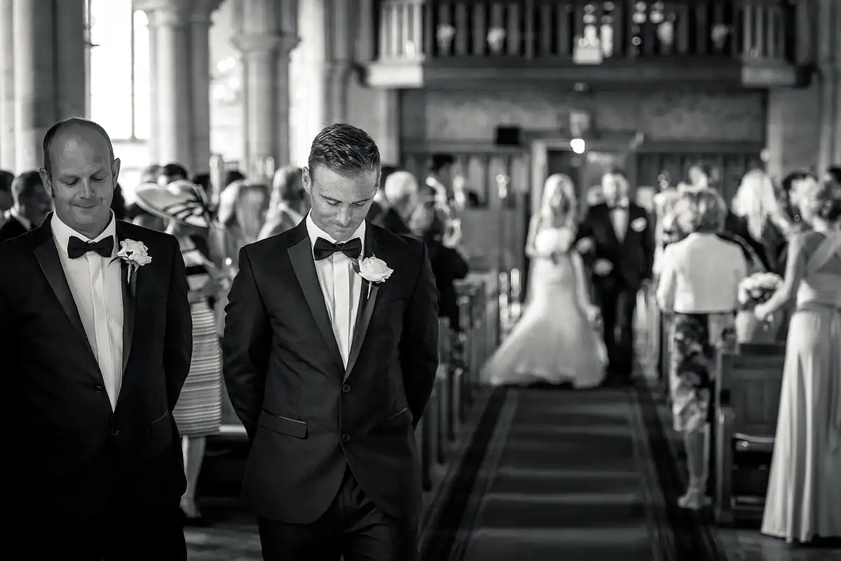 Black and white of the groom and best man in bow ties waiting, heads bowed, as the bride walks up the church aisle behind them.
