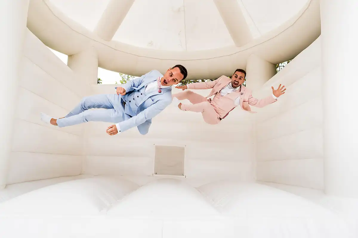 Two grooms in pale blue and pink suits caught mid-air inside a white bouncy castle.