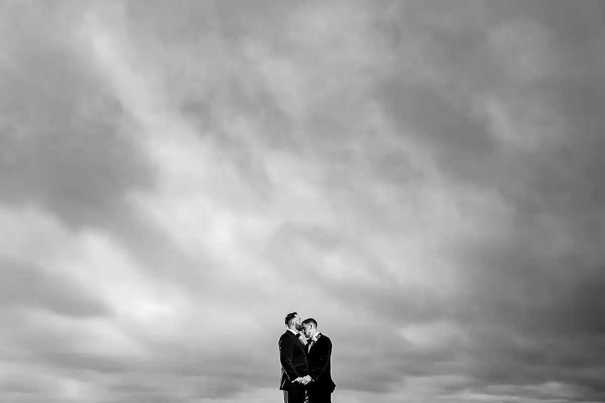 Black and white of two grooms kissing, small at the bottom of a huge cloudy sky.