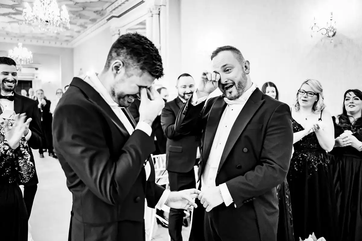 Black and white of two grooms in tuxedos wiping away tears at the end of the aisle as guests smile.