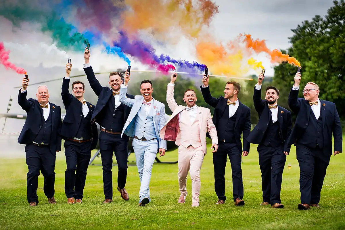 Two grooms in pale blue and pink suits walk with groomsmen holding rainbow smoke bombs overhead on a lawn.