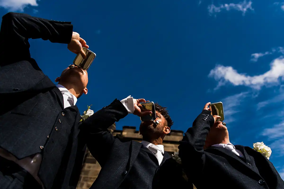 Low view of three groomsmen in dark suits tipping back hip flasks against a deep blue sky.