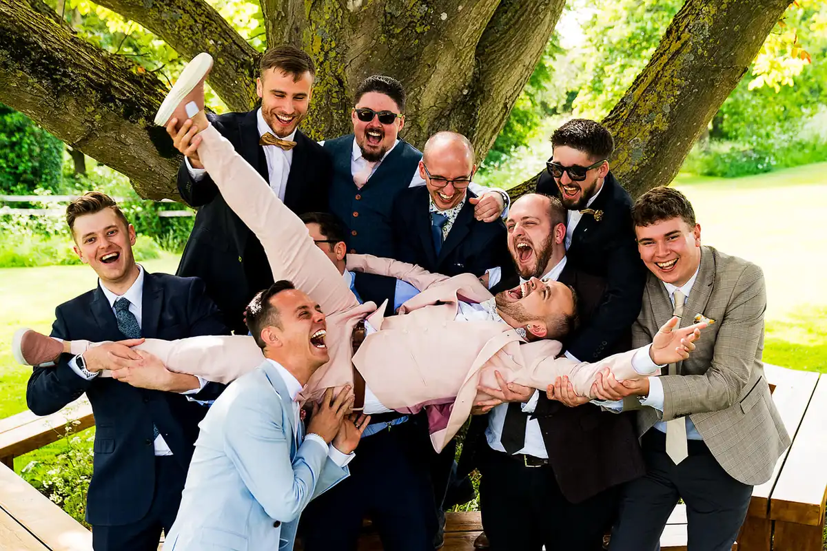 Laughing groomsmen hold a groom in a pink suit horizontally in front of a big old tree.