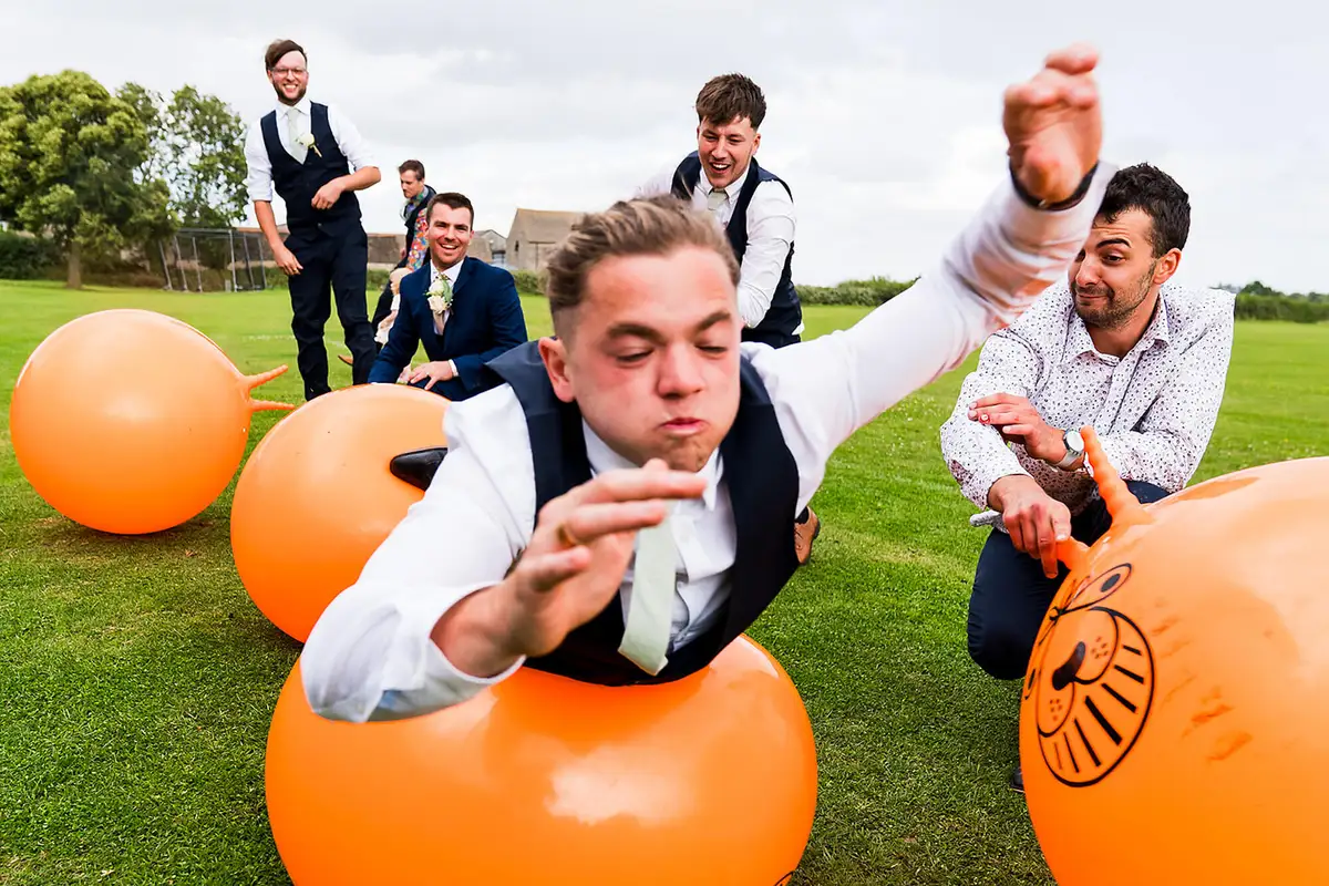 Groomsmen racing on orange space hoppers across a grassy field, one lunging towards the camera.