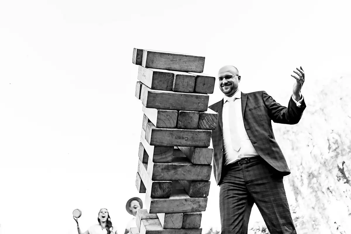 Black and white of a man in a checked suit beside a giant wobbling Jenga tower as guests shriek behind.