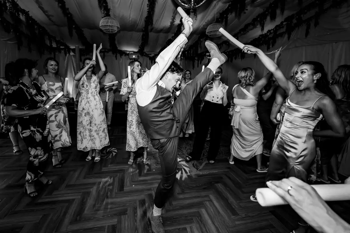 Black and white of a man doing a high kick on the dance floor as guests cheer and wave foam batons.