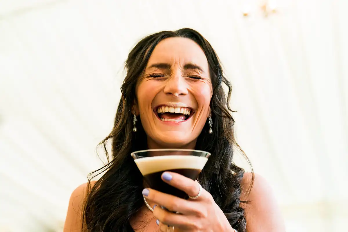 Woman laughing with her eyes shut, holding an espresso martini up to the camera.