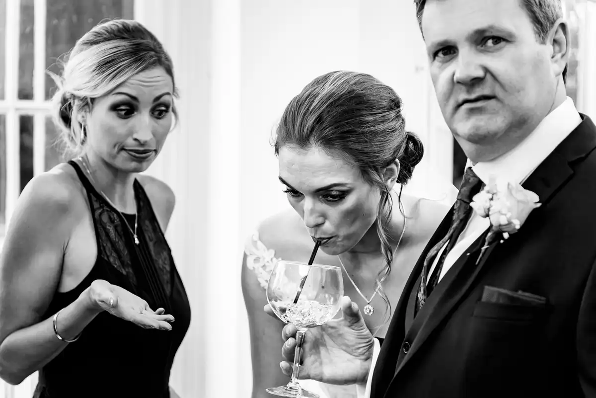 Black and white of a guest shrugging as the bride sips through a straw and a man stares at the camera.