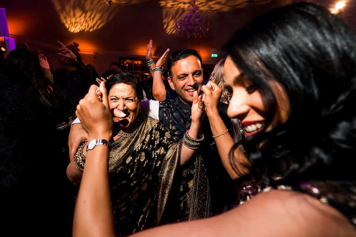 Guests in saris and a patterned shirt dancing and pulling faces under warm patterned party lights.