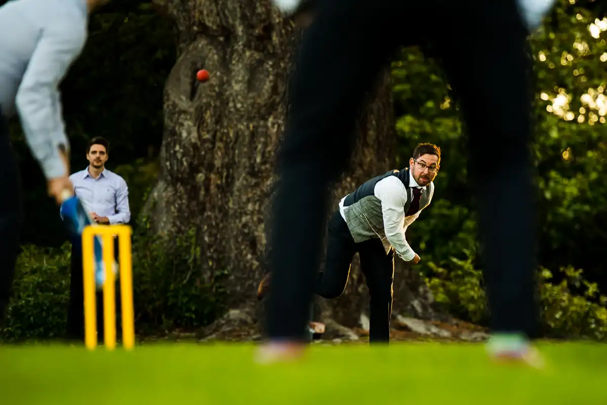 Low view of guests playing cricket on a lawn, the ball in the air as a man in a waistcoat bowls.