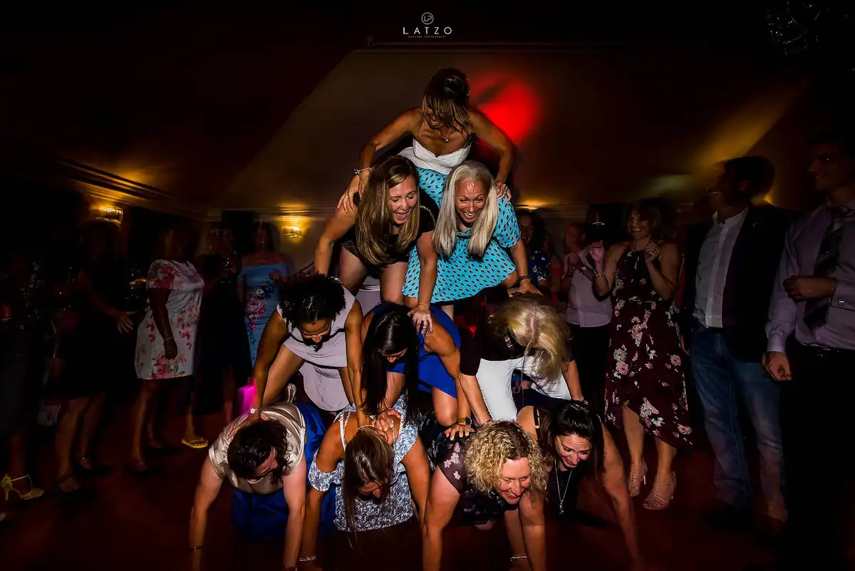 Guests in summer dresses build a wobbly human pyramid on the dance floor, a woman in white on top.