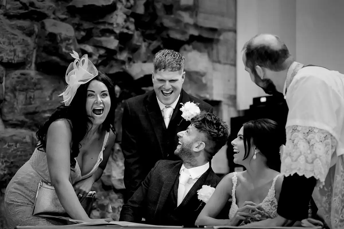 Black and white of the couple signing the register, laughing, as a guest in a fascinator and a groomsman crack up beside them.