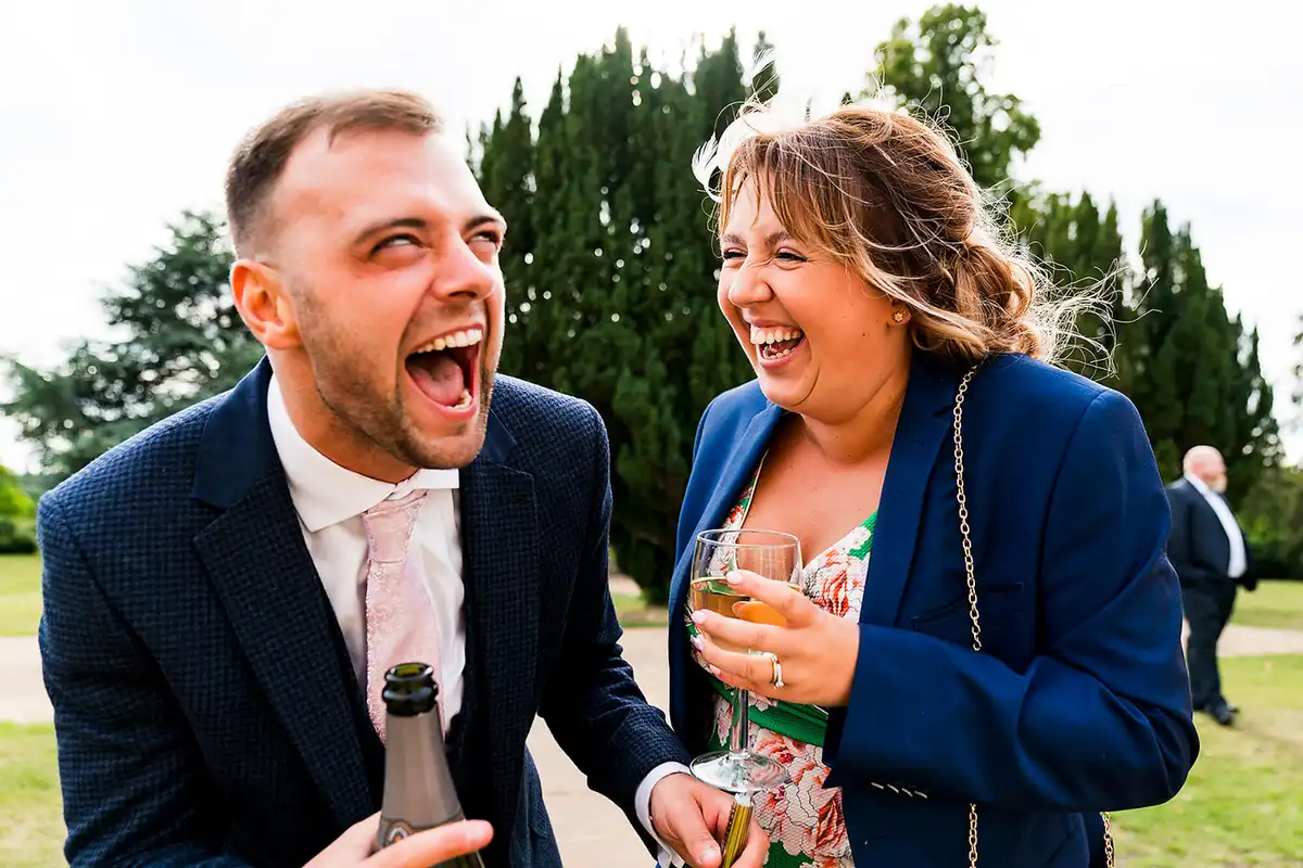 Two guests laughing hard on a garden path, one holding a bottle and the other a glass of fizz.