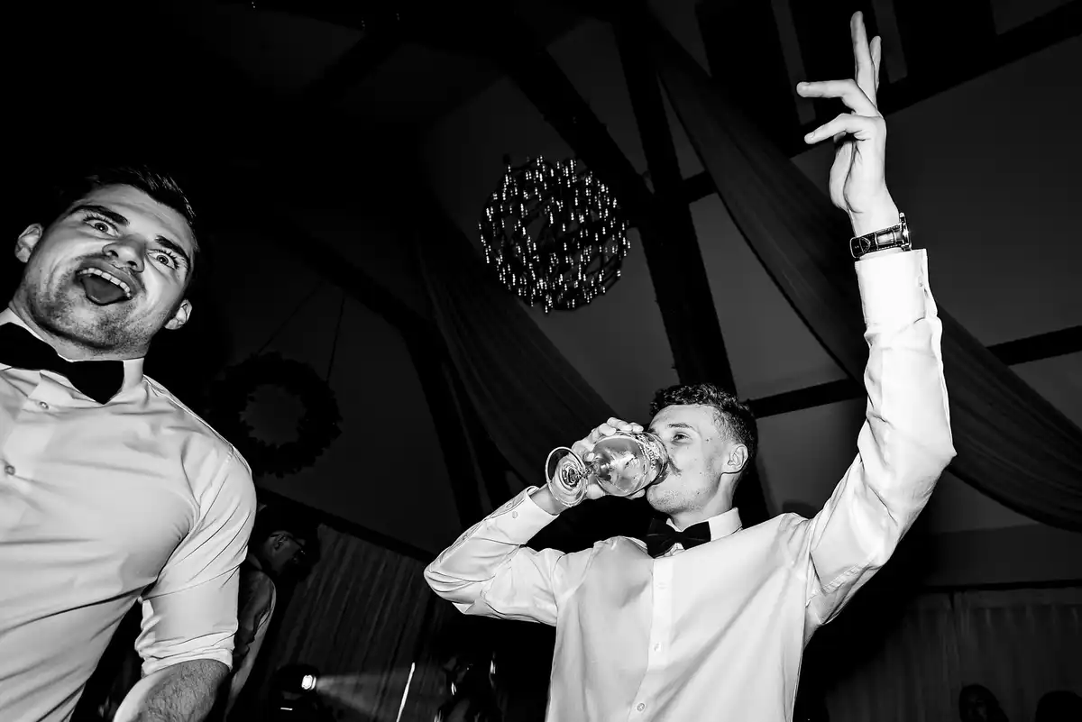 Black and white of two young men in bow ties on the dance floor, one drinking, one pulling a face.