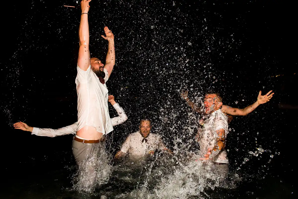 Men in soaked white shirts leap and splash in water at night, spray flying everywhere.