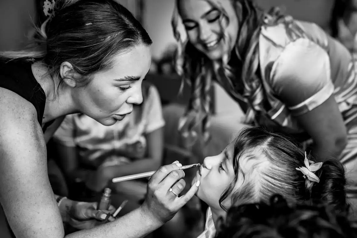 Black and white of a makeup artist brushing lip colour on to a young girl as a bridesmaid watches.