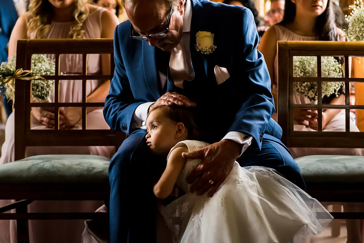 Older man in a blue suit cuddles a sleepy flower girl on his lap during the ceremony, bridesmaids seated behind.
