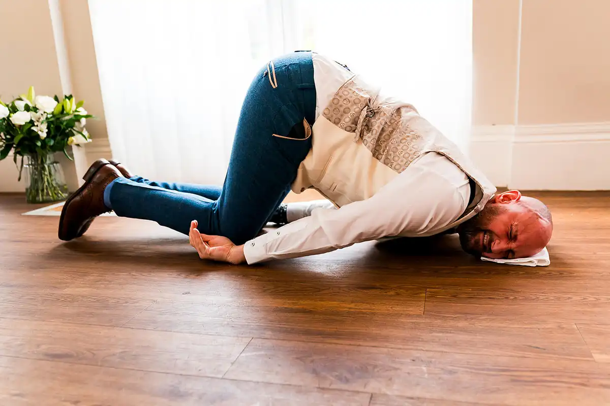 Man in a waistcoat curled up on a wooden floor with his face on a white napkin, grimacing.