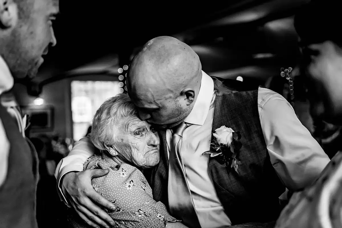 Black and white of a man with a rose buttonhole hugging an older woman tight and kissing her head.