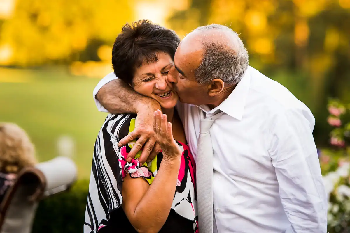 Older man kisses a laughing woman's cheek, arm round her, in warm golden evening light.