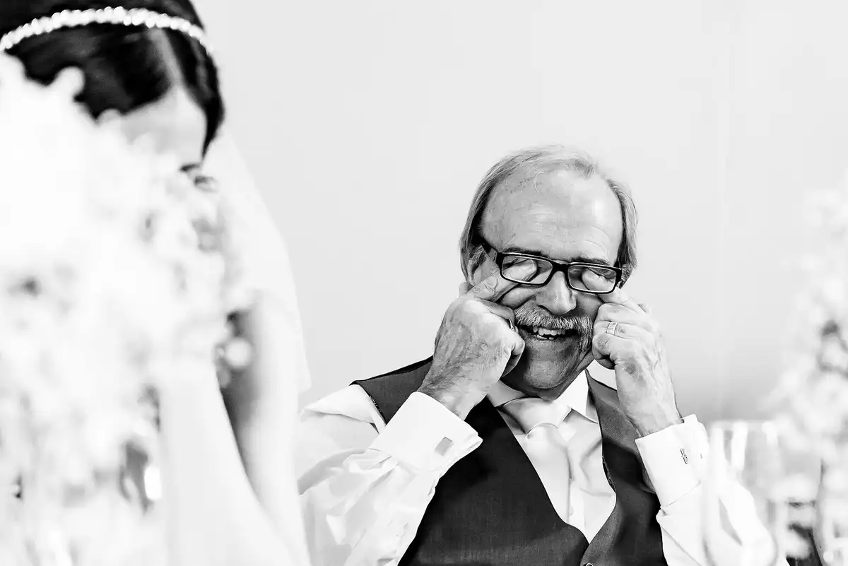 Black and white of an older man at the top table wiping his eyes under his glasses and grinning, the bride blurred beside him.