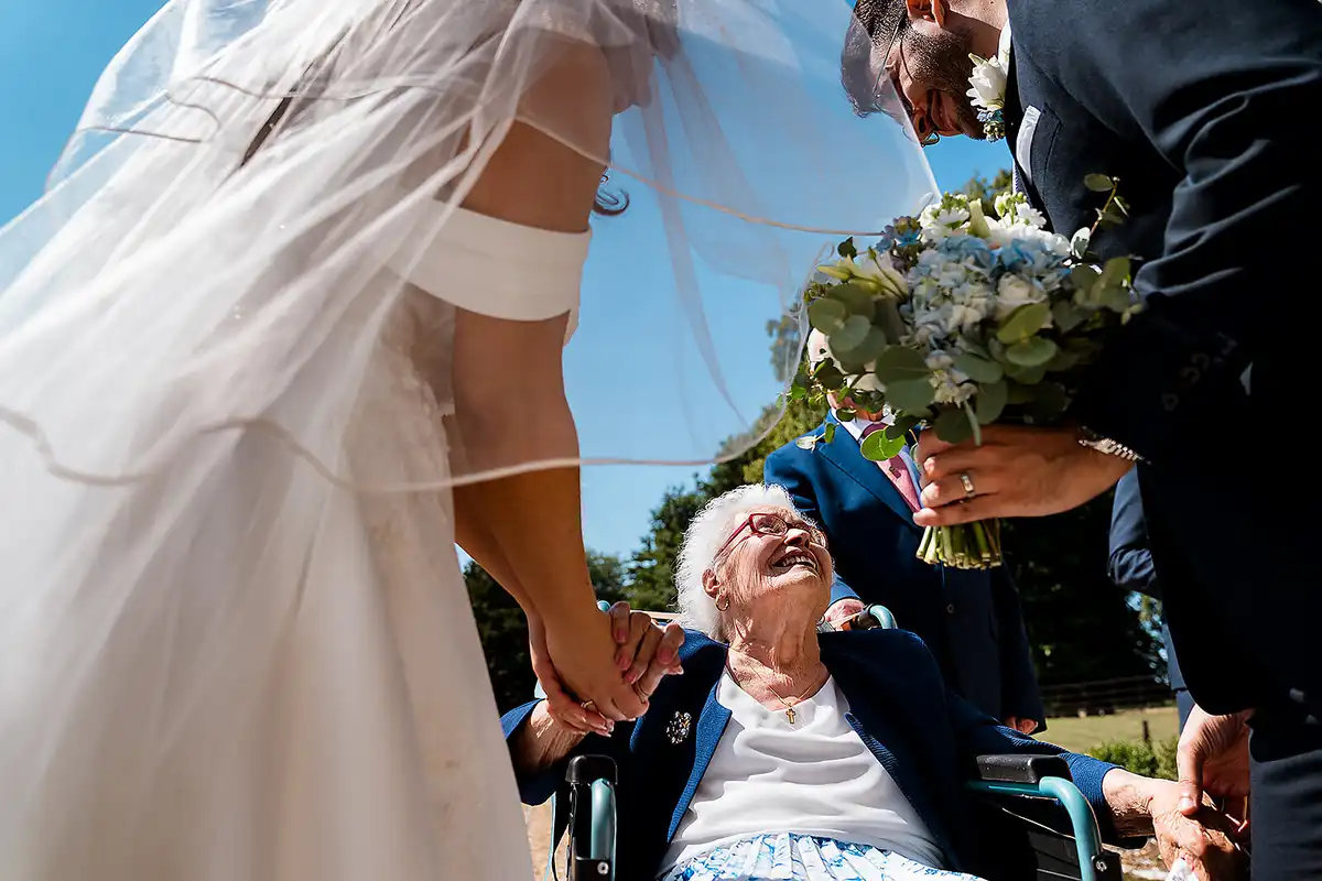 Older woman in a wheelchair laughs up at the bride, holding her hand, the bride's veil blowing across the frame.