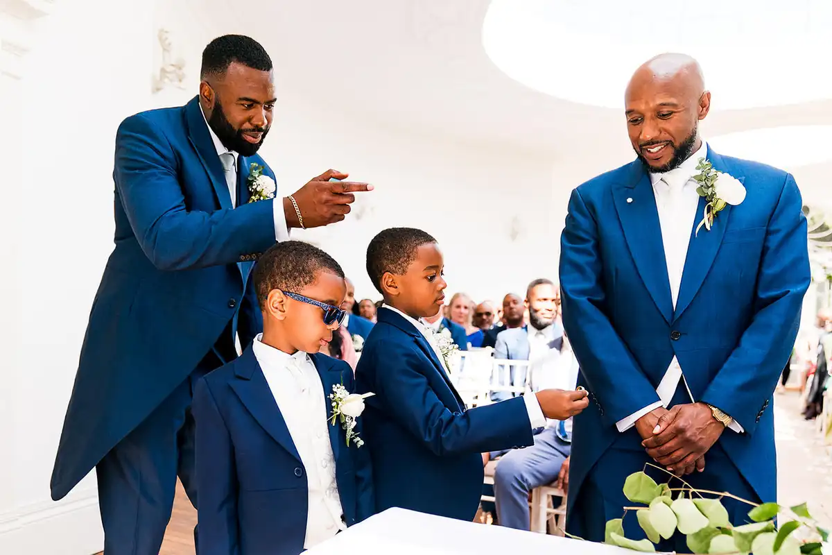 Two page boys in navy suits at the ceremony table as a man in a morning suit points and the groom smiles.