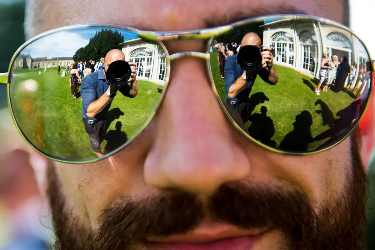 Close-up of a guest's mirrored sunglasses reflecting the photographer and guests on a lawn.
