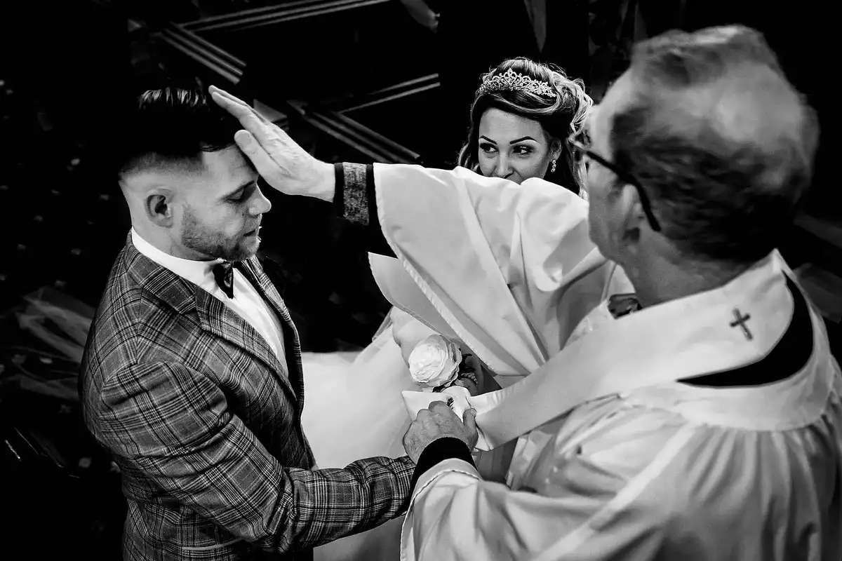 Black and white of a priest with his hand on the groom's forehead as the bride peeks round, laughing.