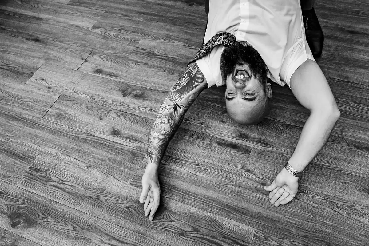 Black and white of a tattooed man lying on his back on a wooden floor, laughing upside down.