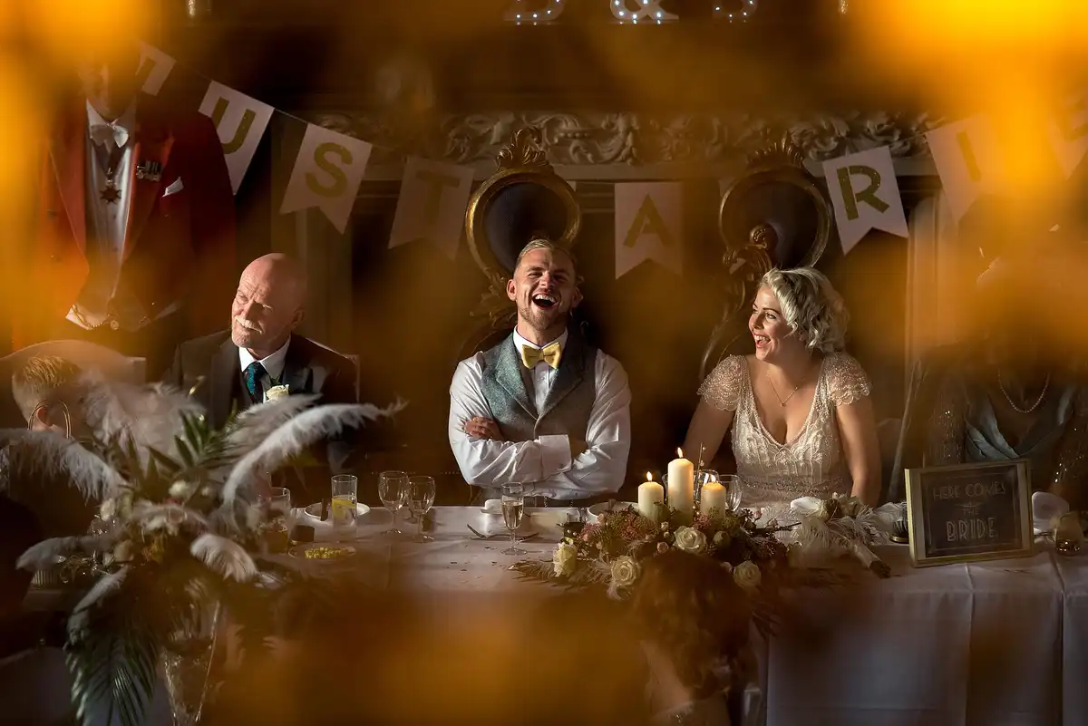 Top table laughing during the speeches, seen through warm blurred foreground with bunting overhead.