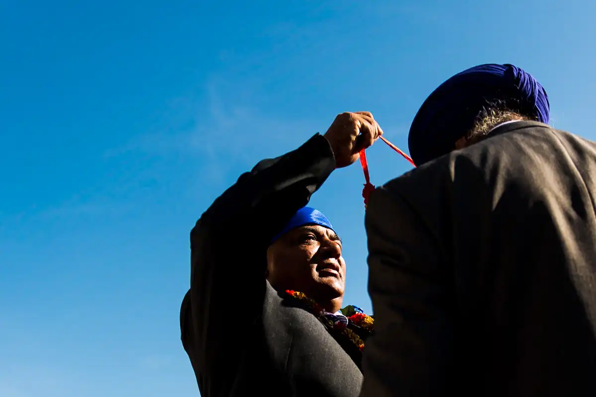 Low view of a man in a blue turban and flower garland holding up a red ribbon beside a man in a purple turban, deep blue sky behind.