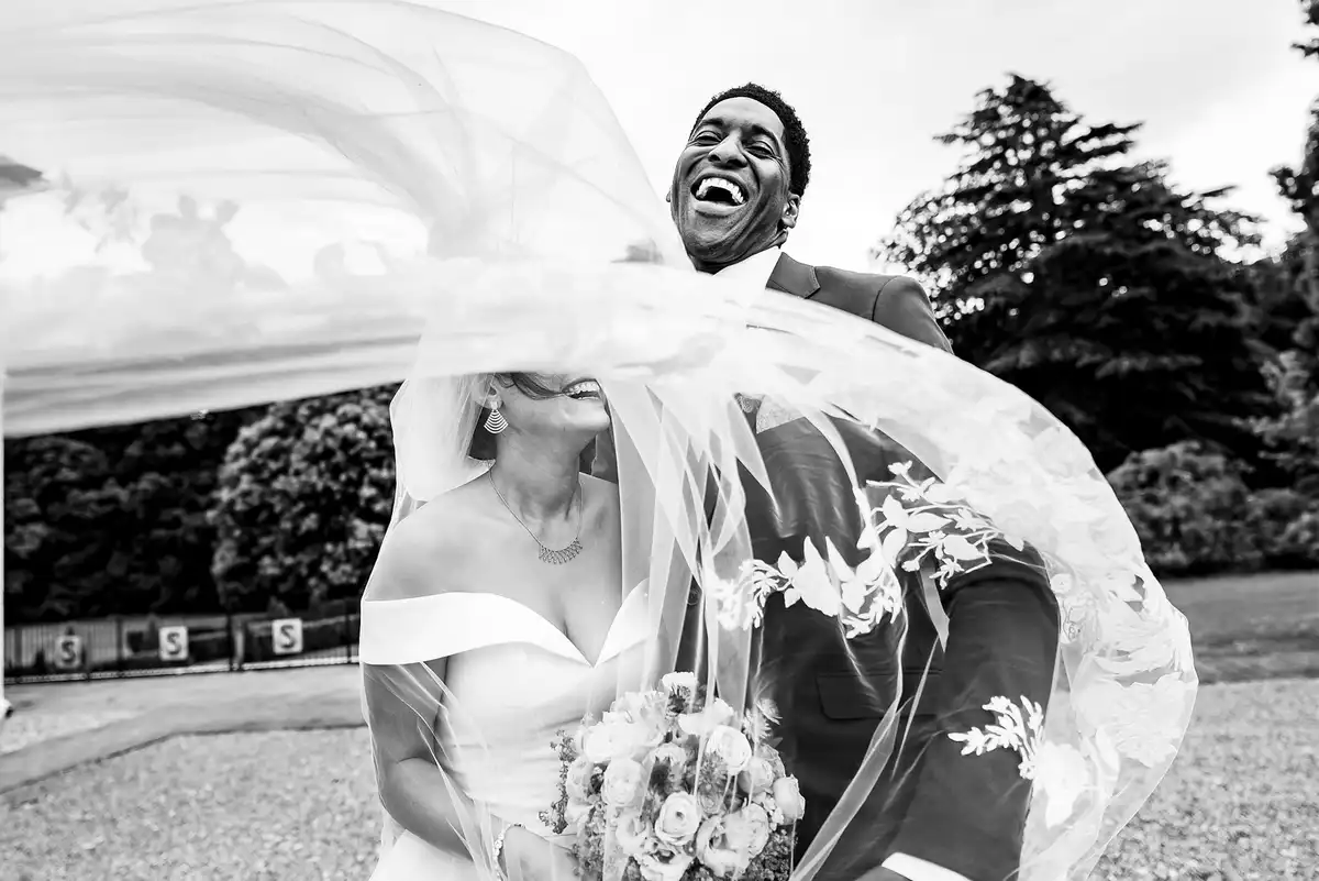Black and white of a lace veil blowing across the bride's face as the groom throws his head back laughing.